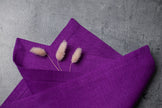 Violet organic linen napkin with mitered corners and dried bunny tail grass on grey stone