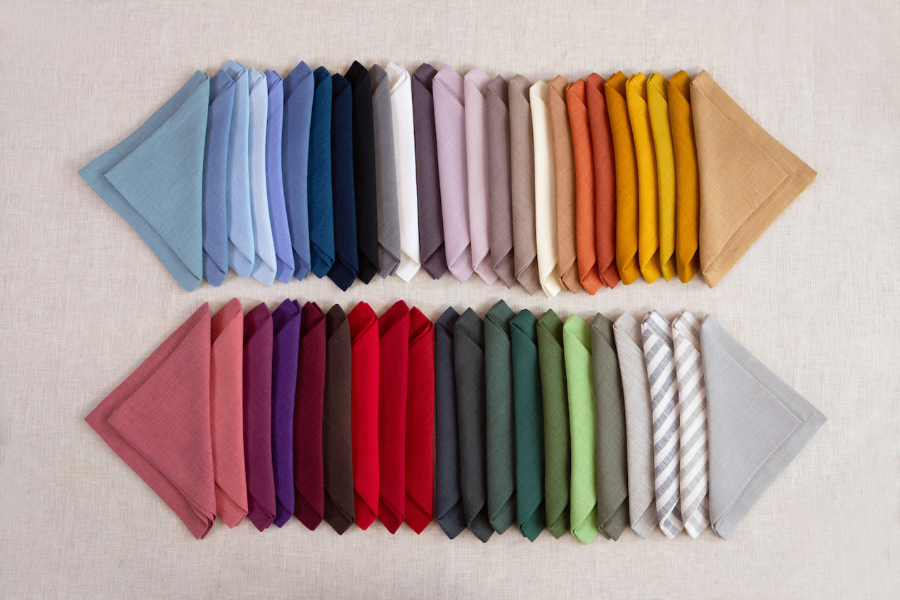 A collection of folded organic linen napkins in various solid colors displayed in two rows with the top row having a light color palette and the bottom row having a dark color palette.