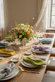 Elegant Easter dining table setting with colorful linen napkins, floral centerpiece, plates, and glasses. Lavender napkin, lime green, mustard, teal an sky blue. 