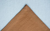 Close-up of warm brown linen fabric with a stitched hem on a pale blue linen background