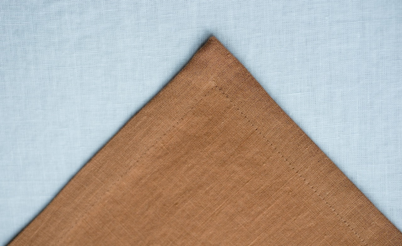 Close-up of warm brown linen fabric with a stitched hem on a pale blue linen background