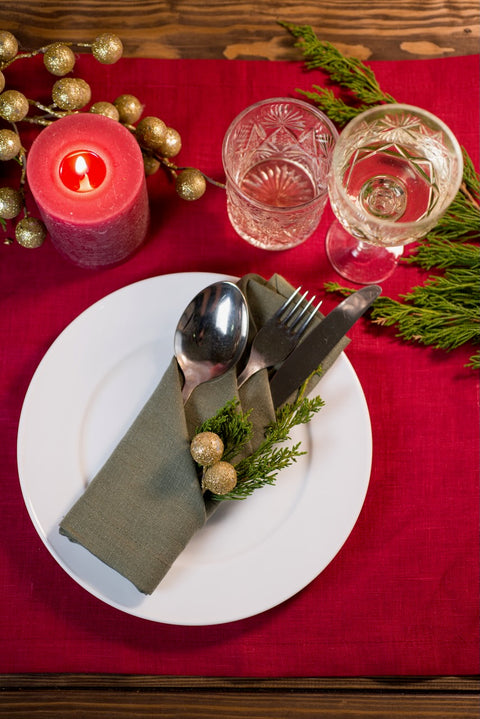 Sage green linen napkin folded into a cutlery pocket with gold berries and evergreen on a red Christmas table