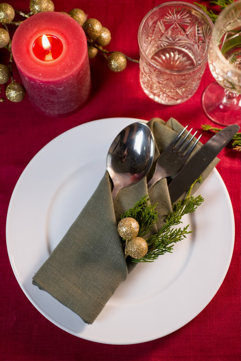 Sage linen napkin cutlery pocket holding spoon, fork and knife next to a red candle and gold baubles