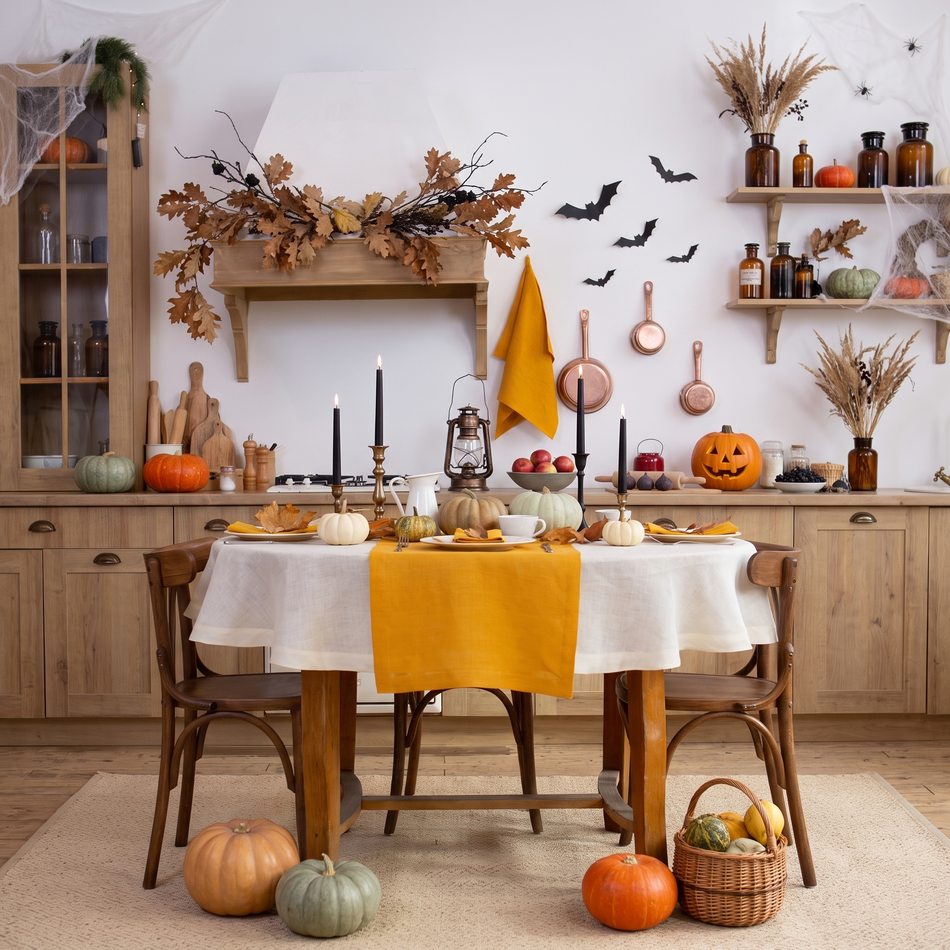 Halloween kitchen table with a natural round linen tablecloth, mustard linen table runner and napkins, black taper candles, pumpkins and paper bats – CoolLinen