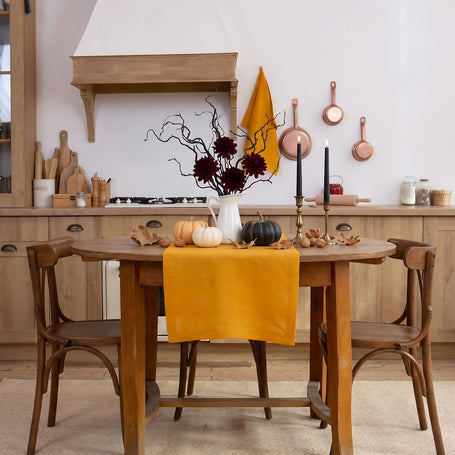 Halloween table with a mustard linen table runner, pumpkins, black taper candles and dark dahlias in a bright rustic kitchen – CoolLinen