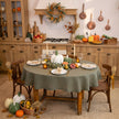 Thanksgiving table with a sage green round linen tablecloth, white and green pumpkins, taper candles and linen napkins in a rustic kitchen – CoolLinen